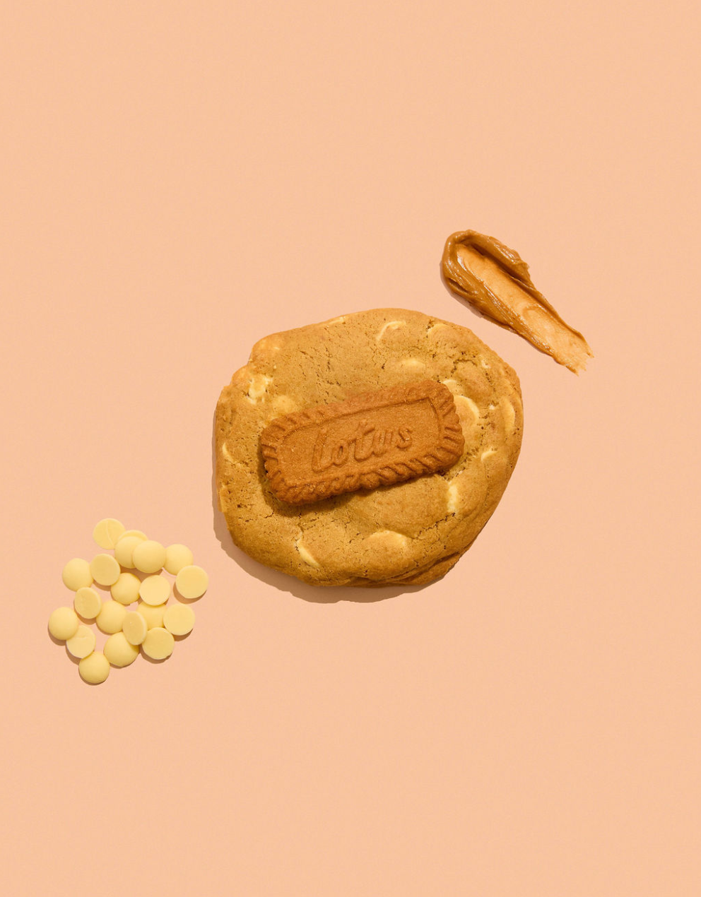 A golden-brown cookie with a embossed 'Lotus' biscuit in the center, styled with a smear of peanut butter and white chocolate chips on a solid peach background.